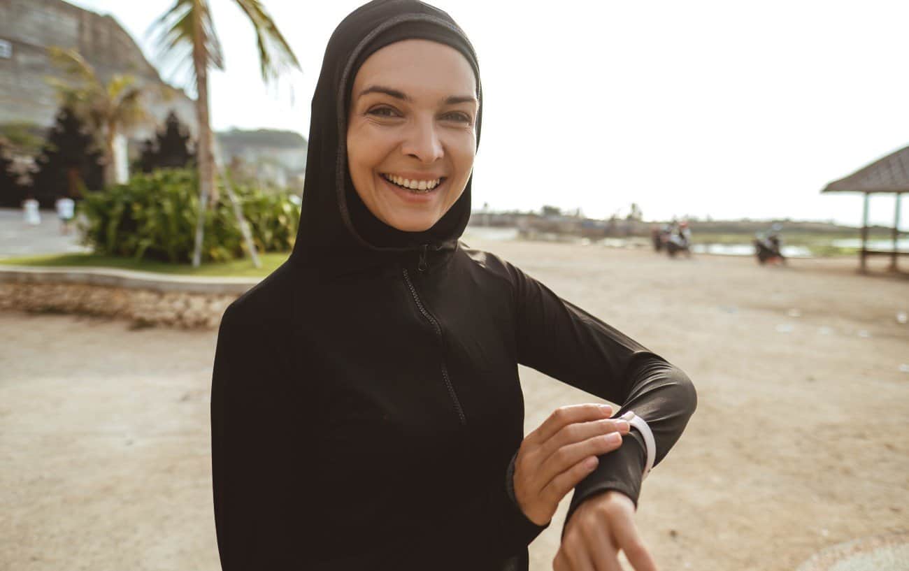 Muslim woman smiling at camera on the beach, checking her GPS watch. 